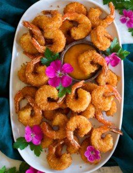 Photo: Many coconut shrimp spread out on a white oval platter with mango sauce in the center in a wooden cup. Vibrant pink flowers are dotted throughout for decoration and platter is resting on a blue cloth over a marble surface.