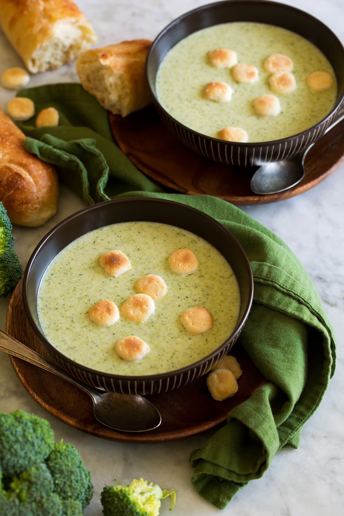 Cream of broccoli soup in brown serving bowls with a crackers atop it. A green cloth is shown to the side and baguette bread as a serving suggestion.