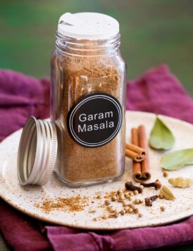 Garam masala spice blend in a glass jar set over a plate with whole spices next to it.