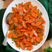Glazed carrots shown served on a white oval platter set over a wooden tray and a green cloth.