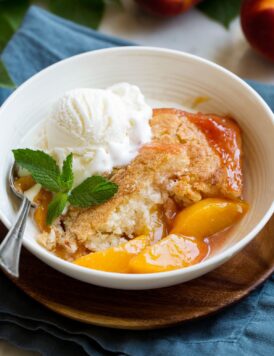 Serving of homemade peach cobbler in a white serving bowl topped with vanilla ice cream and mint.