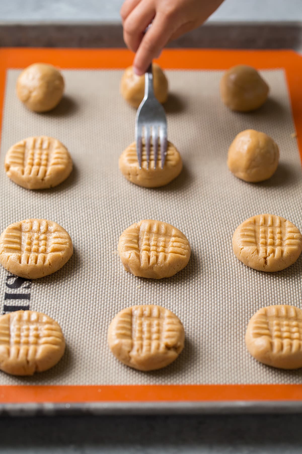 Showing how to press criss cross pattern into peanut butter cookie dough balls on baking sheet.