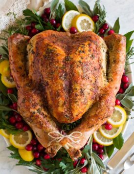 Overhead image of whole turkey on a white platter garnished with fresh herbs, lemon slices and cranberries around it.