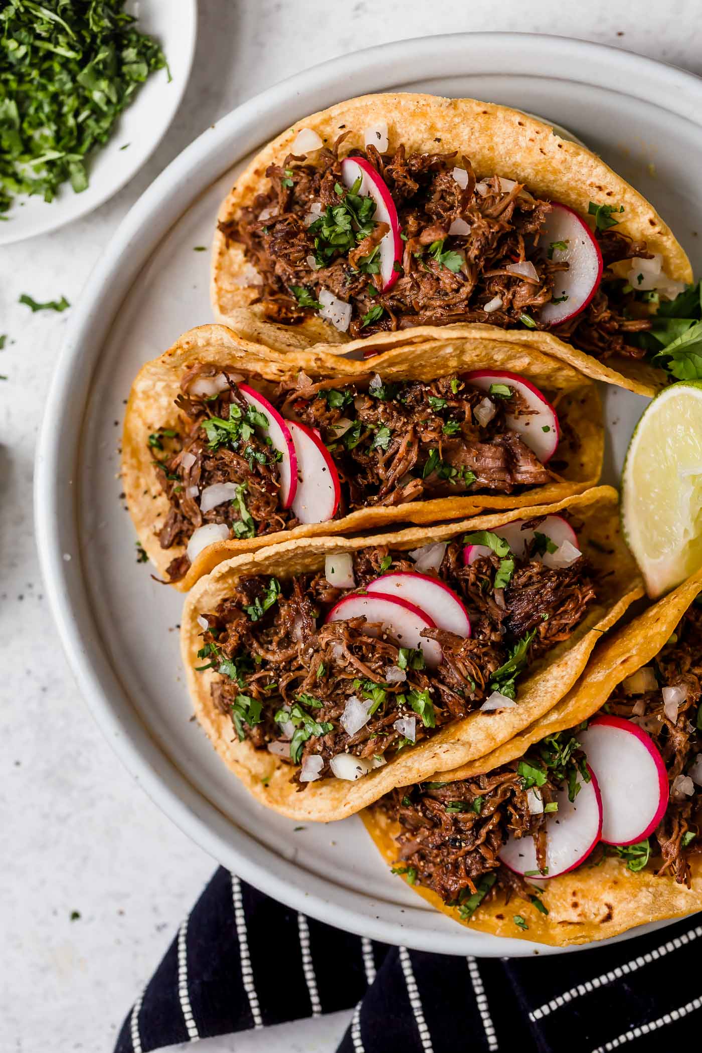 Barbacoa beef layered in corn tortillas to make tacos. Shown sitting on a light grey serving platter, beef is garnished with cilantro and radishes. 
