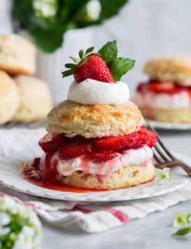 Strawberry shortcake on a white dessert plate. Made up of sweet buttermilk biscuit, macerated strawberries, and whipped cream. It is decorated on top with a fresh strawberry and mint leaves.
