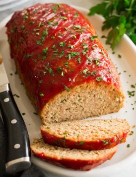 Photo of turkey meatloaf on a white oval serving platter. Parsley is sprinkled over and whole parsley is to the side. A large knife is shown to the side.